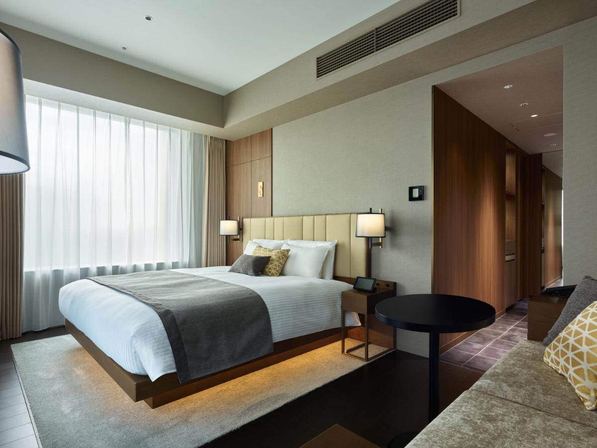 Mitsui Garden Hotel Roppongi Tokyo Premier Rum bild The photo shows a modern hotel room. It features a large bed with a light-colored duvet and decorative pillows. There are bedside tables with lamps on either side of the bed. The room has large windows dressed with sheer curtains, allowing natural li