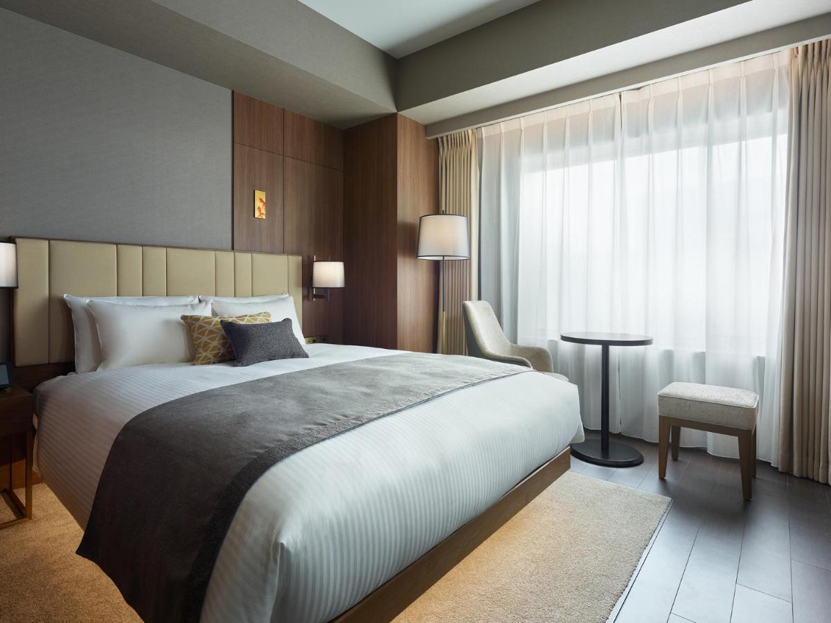 Mitsui Garden Hotel Roppongi Tokyo Premier Exteriör bild The photo shows a modern hotel room featuring a comfortable bed with white linens and a decorative throw pillow. The room has a neutral color palette with wooden accents on the walls. There are two bedside lamps and a chair positioned next to a small