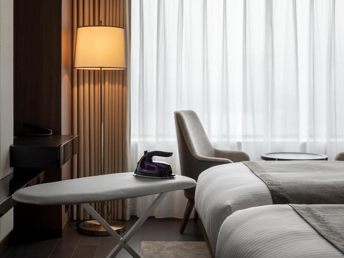 Mitsui Garden Hotel Roppongi Tokyo Premier Exteriör bild The photo shows a modern hotel room featuring two beds with white bedding. There is a light-colored armchair next to a small side table. On the table, there is an iron, and beside it, a lamp with a warm glow is illuminating the space. The large windo