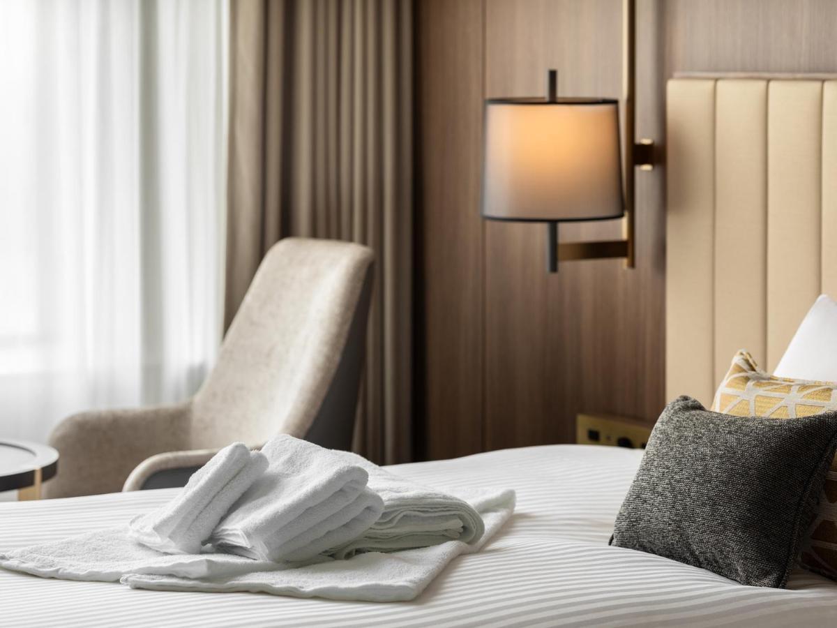 Mitsui Garden Hotel Roppongi Tokyo Premier Exteriör bild The image shows a stylish hotel room. In the foreground, there is a neatly made bed with white linens, featuring a stack of white towels arranged in a decorative manner. To the side, there is a gray upholstered armchair and a small table. The backgro