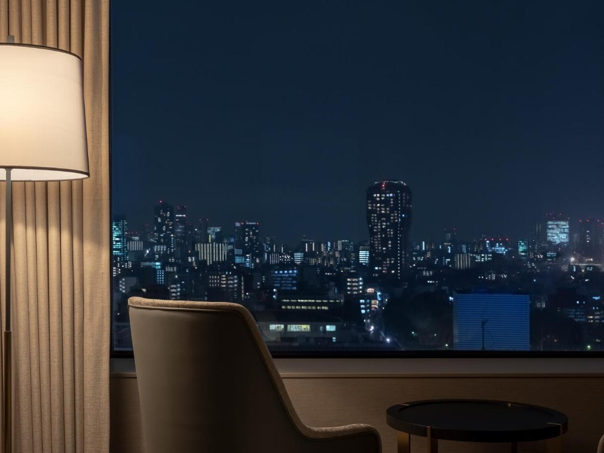 Mitsui Garden Hotel Roppongi Tokyo Premier Exteriör bild The photo depicts a cozy hotel room setting at night. In the foreground, there's a stylish armchair next to a small round table. A standing lamp provides soft lighting, creating a warm ambiance. In the background, large windows reveal a city skyline 