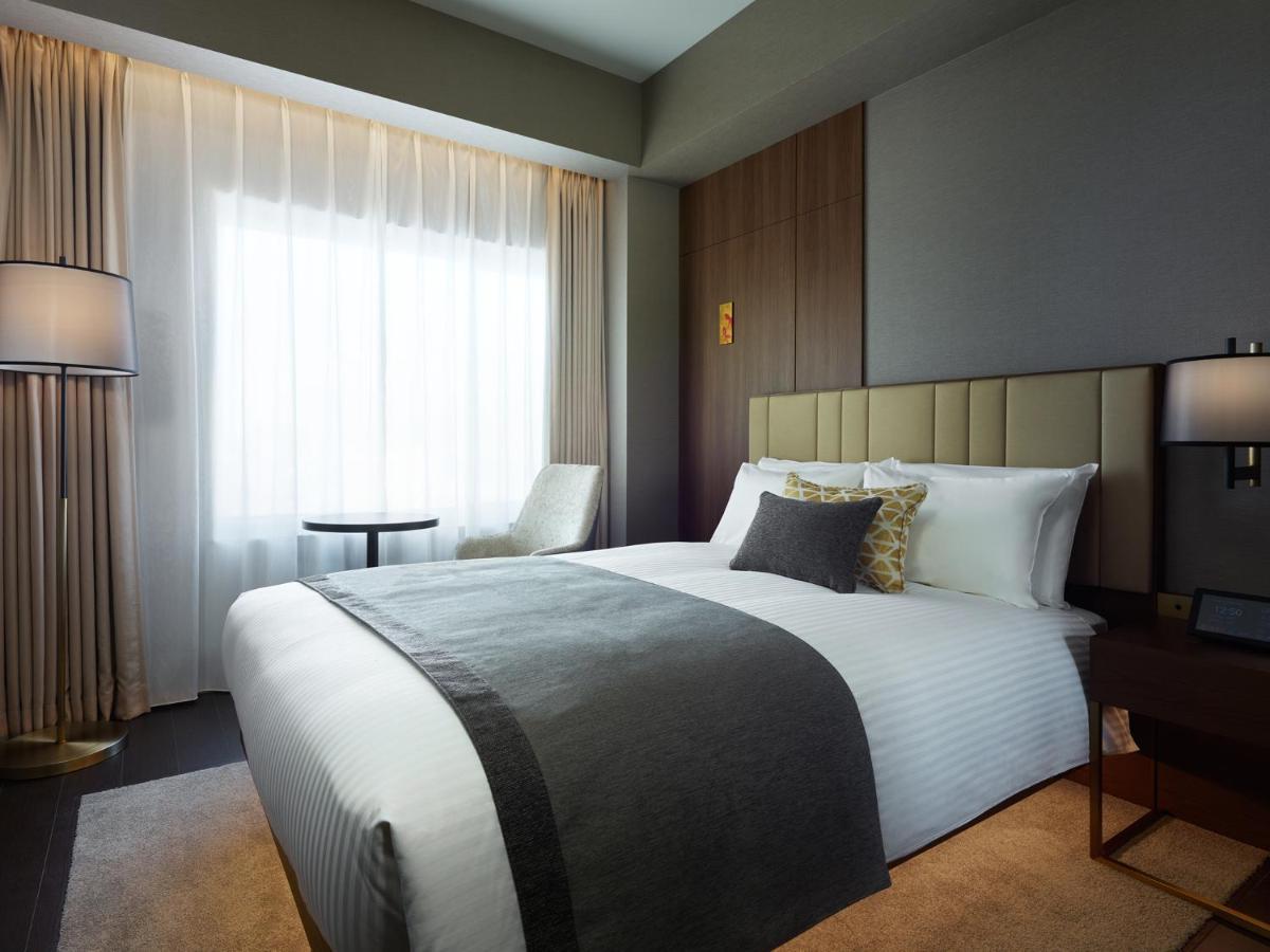 Mitsui Garden Hotel Roppongi Tokyo Premier Exteriör bild The photo shows a modern hotel room featuring a neatly made bed with white linens and a decorative gray throw blanket. To the side, there is a small round table and a comfortable chair, creating a cozy seating area. The room is well-lit with natural 