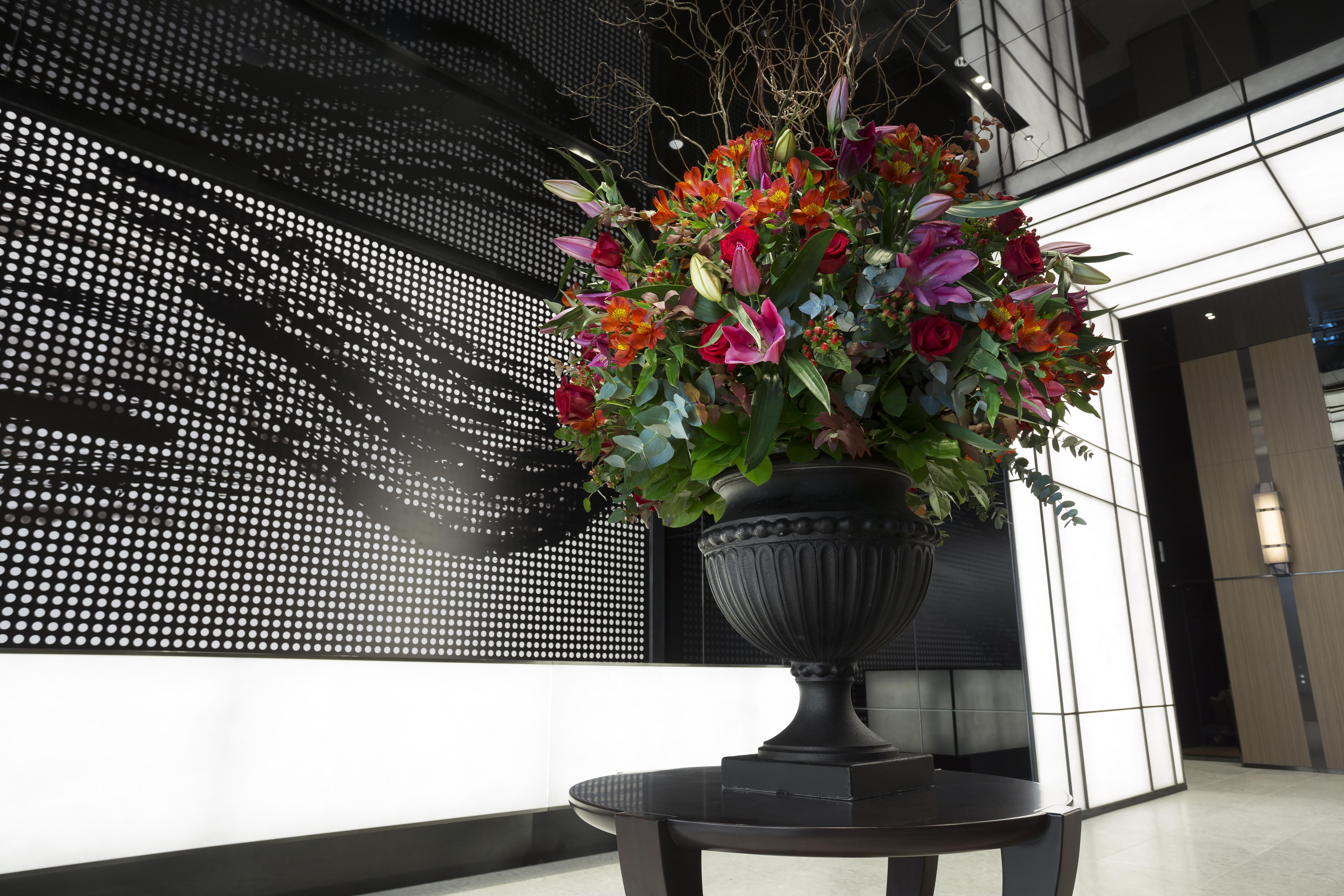 Mitsui Garden Hotel Roppongi Tokyo Premier Exteriör bild The photo features a large, vibrant floral arrangement in an ornate black urn. The arrangement is composed of various colorful flowers and greenery, showcasing shades of red, pink, orange, and green. This floral display is positioned on a small table