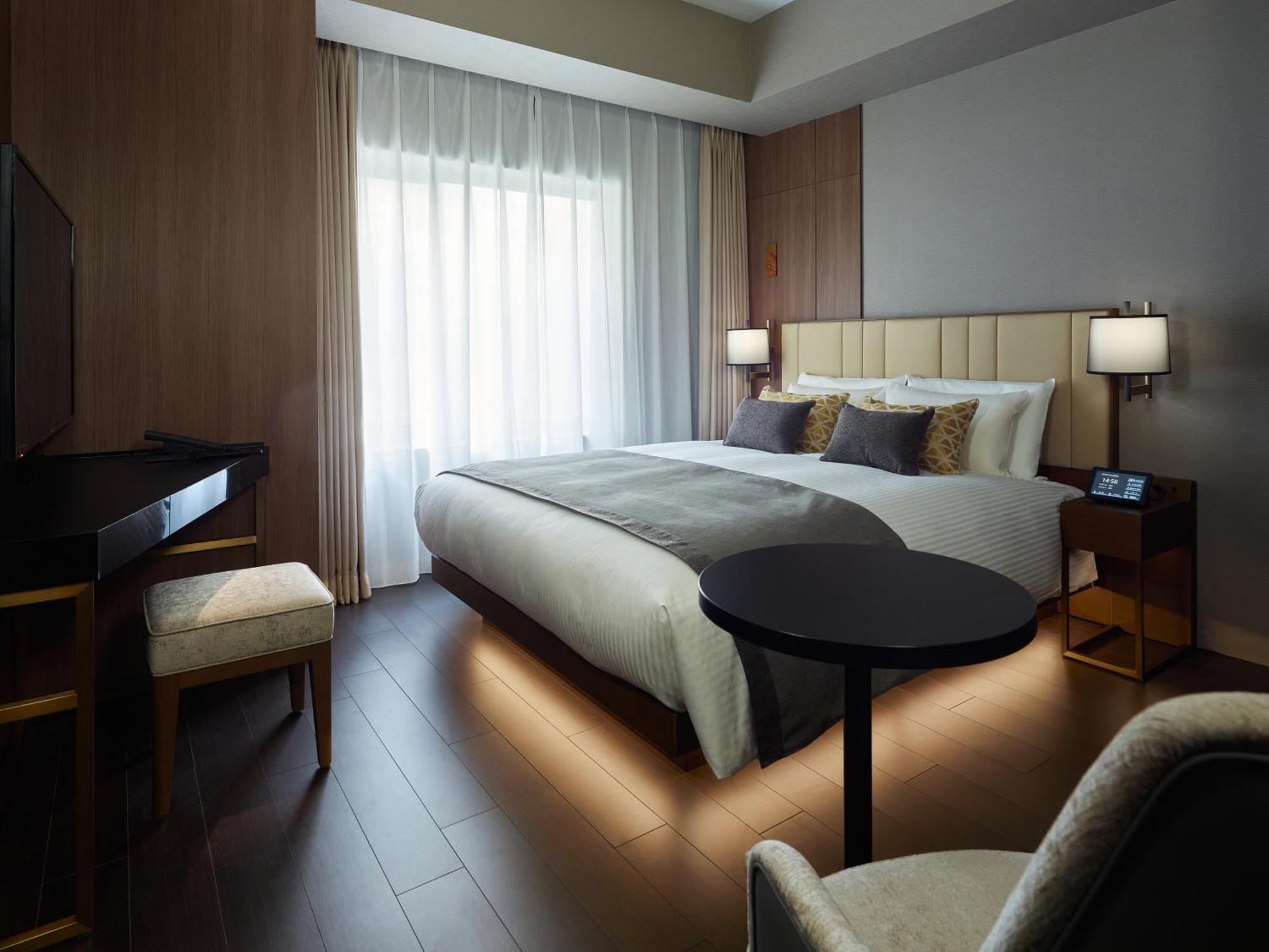 Mitsui Garden Hotel Roppongi Tokyo Premier Exteriör bild The photo shows a modern hotel room featuring a large bed with decorative pillows. There are large windows with sheer curtains allowing natural light to filter in. To one side, there is a sleek desk with a chair and a small round table in the center 