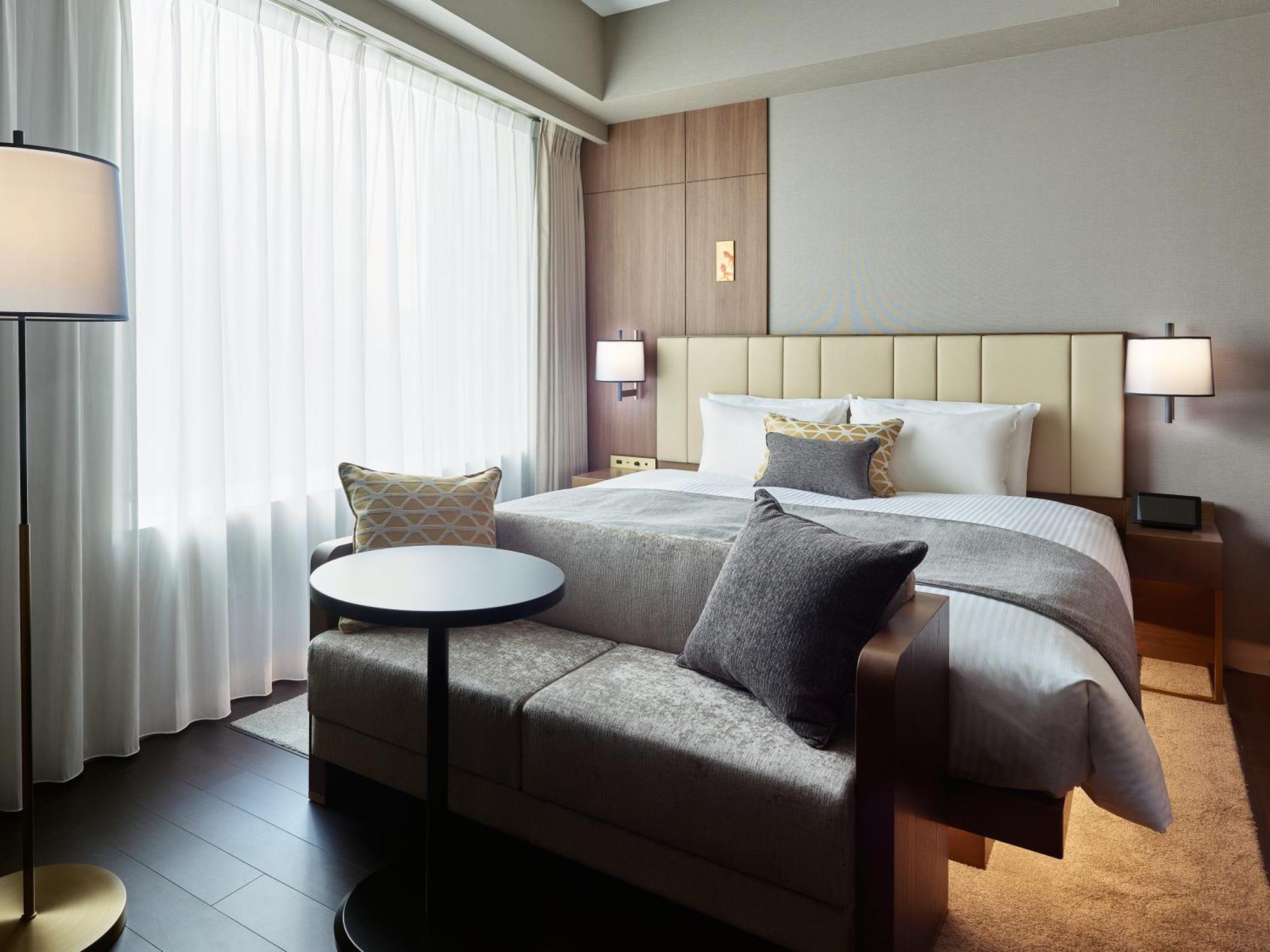 Mitsui Garden Hotel Roppongi Tokyo Premier Exteriör bild The photo shows a modern hotel room that features a large bed with a padded headboard and soft bedding. There's a small, round table accompanied by a couch or seating area, adorned with decorative pillows. To the left, a floor lamp provides additiona